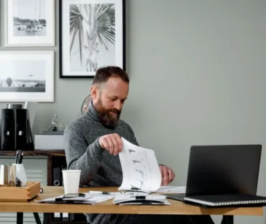 A bearded man reviews documents at a desk with a laptop in a modern office setting.
