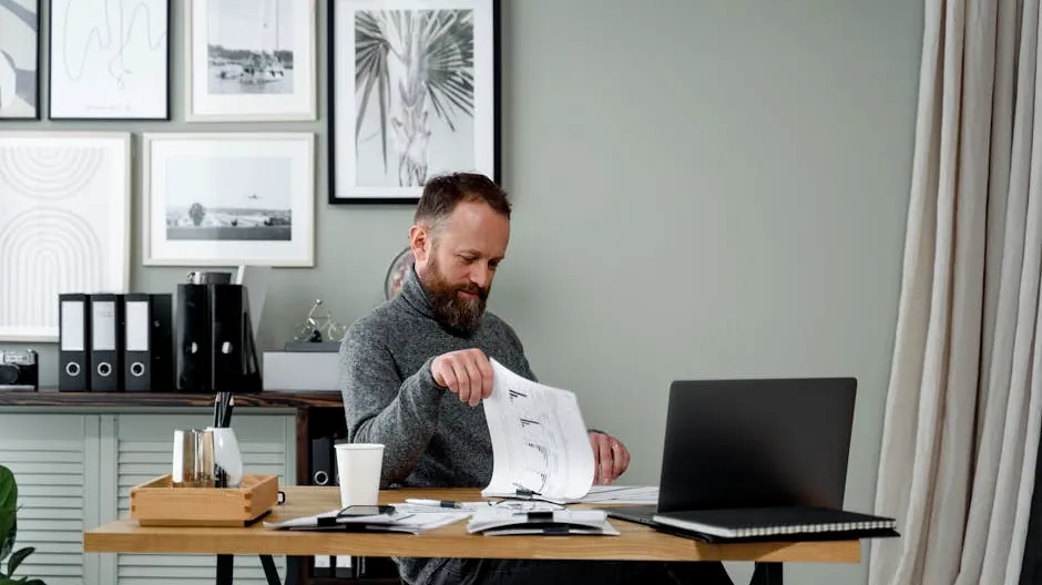 A bearded man reviews documents at a desk with a laptop in a modern office setting.