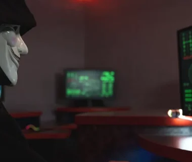 A person wearing a hacker mask operates a computer in a dimly lit room with digital displays.