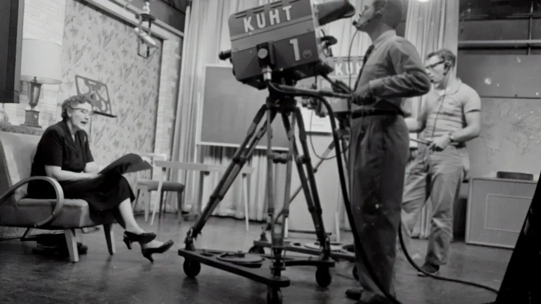 grayscale photography of man standing near studio camera and woman sitting while holding book