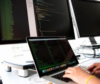 A developer writes code on a laptop in front of multiple monitors in an office setting.