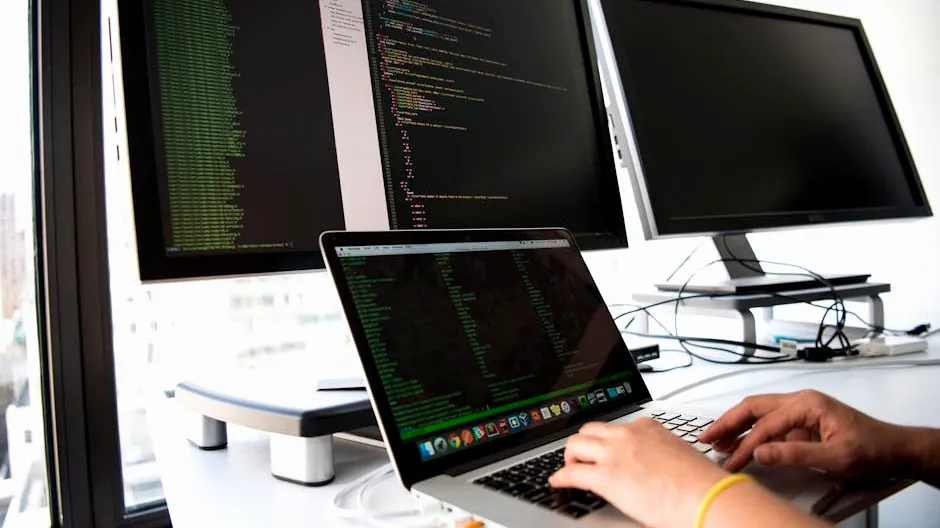 A developer writes code on a laptop in front of multiple monitors in an office setting.