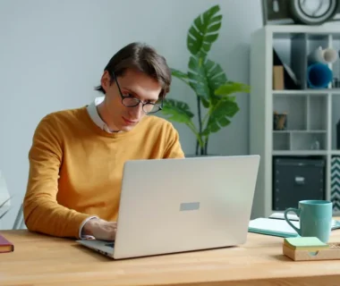 Man in yellow sweater working on laptop at desk.